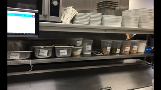A FusionPrep tablet mounted at eye level in a production kitchen, with food prep containers on the shelf below